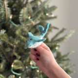 A festive handmade wool felt dolphin finger puppet is held by a hand with dark red nail polish, set against a blurred Christmas tree with white lights. This soft, dual-tone dolphin puppet has a light turquoise body, a white underbelly, fins, a tail, and expressive black eyes, showcasing its playful, handcrafted design.