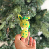 A whimsical green and yellow needle-felted dragon finger puppet is gently held in a woman's hand against a blurred Christmas tree, creating a festive mood. This handcrafted wool felt toy features small green wings, body spots, and tiny ears, perfect for holiday storytelling.