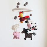 A wool felt farmland animal baby mobile is displayed against a plain white background. It features five wool felt animals: a yellow and burgundy rooster at the top, a white cow with black spots below it, a pink pig on the left, a fluffy white sheep above the pig, and a dark brown donkey in the lower right. All animals hang from a white felt ring, accented with gray felt balls above them.