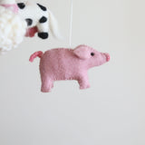 A pink felt pig element from the Farmland Animal felt baby mobile is shown in a close-up, hanging against a stark white background. Crafted from soft pink wool felt, the pig features a small black eye, a snout, and a curly tail with simple stitched details, highlighting its handmade charm. Blurry outlines of sheep and a cow are visible in the background, suggesting other elements of the whimsical mobile.