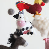 A Farmland Animal Wool Felt Baby Mobile is presented in a close-up against a clean white background, creating a calm and whimsical atmosphere. The central figure is a charming white cow with black spots, a pink snout, and felt horns. Above it, a vibrant red and yellow rooster is visible, while a fluffy white sheep and a blurred donkey are partially seen. A gray felt ball is also visible in the upper left.