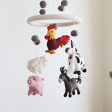 A Farmland Animal wool felt baby mobile hangs suspended against a light, neutral background, creating a calm and whimsical atmosphere. This handmade mobile features plush farm animals: a yellow rooster with red accents, a fluffy white sheep, a light pink pig, a black and white milk cow, and a gray donkey with a black mane. Gray felt balls and a white felt ring complete the mobile, adding balance and visual interest.