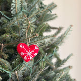 A bright red felt heart-shaped Christmas ornament, part of a set of snowflake Christmas ornaments, hangs by a jute string from a lush green Christmas tree branch. The handmade felt ornament features delicate white snowflake embroidery, contributing to a joyful, festive atmosphere.