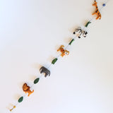 A cheerful wool felt African safari animal garland is displayed diagonally against a bright white background. This needle-felted, 3D felt garland features alternating animals on a thin black string. It showcases an orange lion, gray elephant, striped tiger, zebra, and spotted giraffe with a blue felt ball, ending with a copper bell.