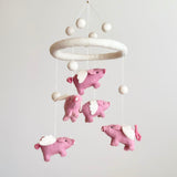 A handcrafted flying pig felt baby mobile is suspended against a plain, light background. Made from soft, natural wool felt, it features a circular felt hoop from which several pink pigs with white wings hang by thin white strings. The charming pigs have detailed curly tails and stitched facial features, creating a whimsical and comforting nursery accent.