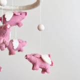 A whimsical wool felt flying pig baby mobile is displayed against a soft white background, evoking a peaceful mood. Several pink wool felt pigs, each with white wings, small black eyes, and curly tails, hang from a white felt hoop by thin strings. A single white wool felt ball completes this charming nursery accessory.