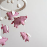A wool felt flying pig baby mobile is shown in a close-up, softly presented against a plain off-white background. The mobile features several handmade natural pink wool felt flying pigs with small white wings, stitched snouts, and black eyes. They are suspended from a circular felt frame, interspersed with small felt balls. The tactile wool felt texture gives a handcrafted feel, suggesting gentle movement for a nursery.
