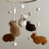 A wool felt baby mobile featuring a Highland Cow theme is presented indoors against a plain background, evoking a calm and playful atmosphere. It features a wooden frame from which soft cotton threads suspend felt Highland cows. The cows, in shades of white, beige, caramel, brown, and chocolate, are crafted with intricate needlework to simulate fluffy fur, suggesting a gentle, comforting rotation.