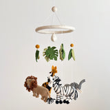 A felt baby mobile, suspended in mid-air against a white background, features a safari jungle theme. The mobile consists of a zebra, giraffe, and elephant, each depicted in a simplistic cartoonish style. The zebra is black and white, the giraffe brown and orange with long necks, and the elephant gray with large ears.