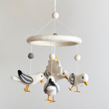 A coastal-themed felt baby mobile with four white felt seagulls is suspended against a light gray backdrop. The charming seagulls feature varied gray and black markings, along with small yellow felt beaks and feet. A white felt ring and assorted white and gray felt balls complete this delicate handmade crib mobile, offering a calm and dreamy atmosphere for a nursery.