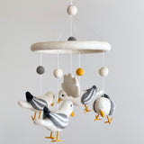 A felt seagull baby mobile is displayed against a plain white background. It features four handcrafted felt seagulls with outstretched wings, predominantly white with grey striped wings and bright yellow beaks and feet. They are suspended from a central ring, above which are white, grey, and yellow felt balls attached by white strings, creating a soothing design.