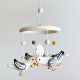 A wool felt seagull baby mobile is displayed against a plain, light gray background. Four charming white felt seagulls with gray markings, yellow beaks, and outstretched wings hang from a circular felt ring. Suspended below are light gray, gray, and yellow wool felt balls, along with two felt cloud shapes, creating a calm and dreamy atmosphere.