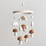 A whimsical felt baby mobile, featuring multiple smiley mushrooms on a beige background, suspended from the ceiling. The mobile is crafted with wool felt and boasts five adorable smiley mushrooms in beige, attached to a white frame with small clouds as decorative elements.