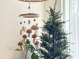A cozy indoor setting featuring a felt baby mobile hanging from the ceiling, showcasing a white wool felt frame with five mushroom-shaped elements each bearing a smiling face. The background includes a Christmas tree adorned with ornaments and lights, suggesting a festive atmosphere.