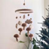 A charming felt baby mobile, 'Felt Baby Mobile - Smiley Mushroom', hangs in front of a Christmas tree. The white frame is adorned with five beige smiley mushrooms, adding a touch of magic to the scene.