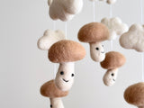 A whimsical felt baby mobile, suspended from the ceiling, features five unique beige mushrooms of varying sizes and shapes. The largest mushroom serves as the centerpiece, surrounded by smaller ones, creating an interesting visual dynamic. Crafted on a white wool felt frame, this handmade mobile adds a touch of warmth and comfort to any nursery.