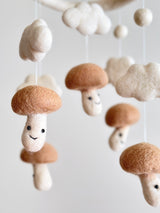 A whimsical felt baby mobile featuring five smiling mushrooms on beige tones attached to a white felt frame, perfect for a child's nursery. The playful design creates a nature-inspired environment with a touch of fantasy and forest magic.