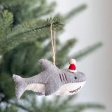 A wool felt baby shark ornament, complete with a red Santa hat, dangles from a Christmas tree branch. This charming felt baby shark ornament features a light gray upper body, a contrasting white lower section with stitched teeth, and a fluffy white pompom on its hat. A jute string allows it to hang playfully, adding a unique touch to festive decor.