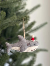 A delightful wool felt baby shark ornament is prominently displayed on a vibrant green Christmas tree. This gray and white shark features a festive red Santa hat with a white pompom. Its charming details include three stitched gills, prominent teeth, a black stitched eye, and a black stitched mouth. A jute string is attached, ready to hang this unique Christmas ornament.