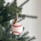 A handmade wool felt baseball ornament hangs on a Christmas tree, creating a festive, sporty atmosphere. The white ornament features red stitching, closely resembling a real baseball. It is suspended by a rustic jute string with a small tag, against a blurred green tree and white wall.