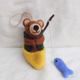 A whimsical needle felted bear fishing ornament is depicted sitting in a yellow boat against a bright white background. This charming brown felt bear wears a green vest and holds a brown fishing rod with a white string leading to a small blue felt fish. A jute string for hanging is attached to the bear.
