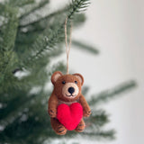 A handcrafted felt bear holding heart ornament is suspended from a green pine branch. This brown felt bear has a lighter tan muzzle and black eyes. It clutches a bright red heart to its chest, ready to hang by its jute string.
