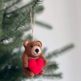 A handcrafted felt bear holding heart ornament hangs from a Christmas tree branch, suspended by a jute string. This brown felt bear ornament features a lighter face, inner ears, and black eyes, holding a small red heart. Its textured surface highlights its charming handmade quality.