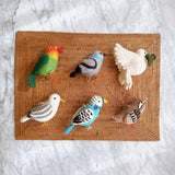A whimsical set of six wool felt bird ornaments, including a vibrant rainbow parrot, grey and pink pigeon, white dove, grey seagull, blue budgerigar, and white throat sparrow, is playfully arranged on a woven mat against a marble backdrop. Each handmade ornament features wire legs; the white dove charmingly holds a green twig.