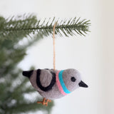 A wool felt pigeon ornament, part of a felt bird ornament set, hangs serenely from a pine branch. This charming gray felt pigeon ornament is crafted from 100% wool felt, featuring a black-tipped head and tail, a black curved side mark, and pink and blue bands around its neck. Brown wire legs and a jute string complete this unique Christmas tree ornament.