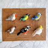 Six charming wool felt bird Christmas ornaments are arranged on a brown woven rattan placemat, which rests on a white marble surface. This set of handcrafted felt ornaments includes a robin, green canary, blue tit, sparrow, woodpecker, and white parrot, each featuring intricate needle-felted details and wire legs. Each bird boasts unique coloring, like the robin's grey and red, the canary's green, yellow, and light blue, and the woodpecker's black, white, and red.