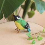 A vibrant felt bird ornament rests on a light wooden round adorned with green leaves, set against lush greenery. This handcrafted wool felt Christmas ornament features a black head with a white marking, a bright green back with white dashes, a yellow lower body, and a blue tail. Delicate wire legs and a red string enhance its realistic detail, making it a charming addition to any holiday decor.
