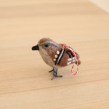 A charming needle-felted wool felt sparrow ornament, part of a set of six felt bird ornaments, rests on a light-colored wooden surface. This realistic sparrow features a plump body of brown, grey, and white wool felt, with black and white markings on its wings. It has a small black beak, delicate wire legs, and a red and white cotton hanging string.
