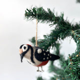 A charming wool felt woodpecker ornament, part of a festive bird ornament set, hangs gracefully from a Christmas tree branch against a clean white backdrop. This handcrafted felt woodpecker ornament features a vibrant mix of white, black, red, and tan wool with needle-felted details. Distinctive features include a red patch on its head, black stripes on its sides, and wire legs.