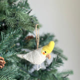 A handmade needle felted white parrot ornament, from a set of unique Christmas felt bird ornaments, is showcased hanging on a vibrant green Christmas tree. The white parrot features a predominantly white body with a bright yellow crest, a small light brown beak, and a touch of orange near its eye. It hangs gracefully by a jute string among the pine needles, next to a natural pine cone.