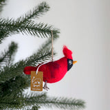 A handmade felt cardinal ornament hangs from a green Christmas tree branch. This festive wool felt cardinal ornament is predominantly red with black facial markings and a small yellow beak. It features a tufted crest and realistic wire legs, ready to hang by its jute string.