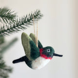 A handcrafted wool felt hummingbird ornament hangs from a deep green Christmas tree branch, set against a soft, neutral backdrop. The needle-felted hummingbird features light green wings with darker accents, a white front, and a dark green head with red markings. A slender black beak extends from its face, and a jute string is attached for easy hanging.