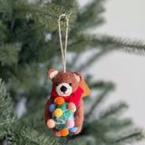 A charming felt brown bear holding a Christmas tree ornament hangs on a green Christmas tree branch. This handmade wool felt bear wears a red scarf and holds a green Christmas tree adorned with colorful bulbs. The unique Christmas tree ornament features a brown bear with rounded ears, a black nose, and black eyes, with a jute string for hanging.