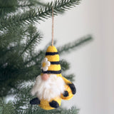 A handcrafted needle felted bumblebee gnome ornament hangs on a pine tree branch, suspended by a thin jute string, against a bright background. This unique Christmas ornament features a conical hat with yellow and black stripes, adorned with a small white flower. A fluffy white beard extends from beneath the hat, and the gnome holds a small, rounded beehive.