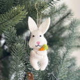 A charming white felt bunny ornament hangs against a soft, blurred green background. This handmade felt bunny ornament features long ears, black eyes, a pink nose, and a stitched black mouth. It holds a small, vibrant felt carrot with green leafy tops, and has a jute string for hanging.