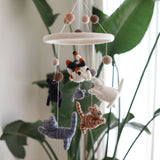 A playful Felt Cat Baby Mobile hangs in a softly lit, natural setting, with a blurred green houseplant and sheer white curtains in the background. Six adorable felt kittens—white, black, gray, tabby, orange tabby, and calico—along with a small gray felt mouse, dangle from a circular white felt ring on thin strings. Small brown fuzzy balls also adorn the strings, creating a comforting and visually engaging nursery accessory.