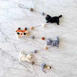 A handmade wool felt cat garland is displayed on a white marble surface. It features four felted cats in black, white, grey, and orange, strung together with small felt balls in muted earth tones. A copper bell hangs at the end of the garland.