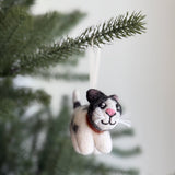 A Calico felt cat ornament hangs on a Christmas tree branch against a soft white background. This handcrafted felt cat is made from 100% New Zealand wool, featuring a white body with gray and black markings, a pink nose, a stitched smile, and white whiskers. A white string is attached for hanging, adding a whimsical touch.