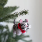 A handcrafted wool felt gray tabby cat ornament is suspended from a fir tree branch, creating a festive display. This unique Christmas ornament, made from 100% New Zealand wool, showcases a gray tabby cat with distinct gray and darker gray stripes on its body and tail. A cheerful red scarf completes the cat's look.