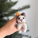 A felt Calico cat ornament is gently held by a female hand against a soft, green-tinted background. This handcrafted wool felt ornament features a blend of white, orange, and black felt, detailed with a pink nose, stitched whiskers, tiny black eyes, and small rounded paws. A delicate tan string is looped around its neck.