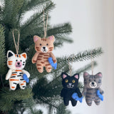 Multiple wool felt cat ornaments, each a stitched cat holding a fish, hang from the green branches of a Christmas tree against a bright white background. These handcrafted cat ornaments feature intricate hand-stitched feline details. Visible variations include a white cat with black and orange stripes, a light brown cat with dark brown stripes, a black cat, and a gray cat with dark gray stripes. Each charming cat holds a small blue felt fish and hangs by a jute string.