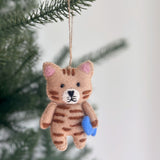 A playful wool felt cat ornament, a stitched cat holding a blue fish, hangs from a jute string against a blurred pine branch and white wall. This light brown cat features darker stripes, pink inner ears, a white muzzle, and intricately stitched edges, evoking a cozy feel perfect for Christmas tree decorating.