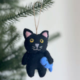 A hand-stitched black felt cat ornament, holding a blue felt fish, hangs on a softly lit Christmas tree. This charming wool felt cat ornament features pink inner ears, yellow eyes, a gray nose, and embroidered white whiskers. A jute string is attached for hanging.