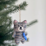 A charming wool felt cat Christmas ornament, handcrafted with a stitched tabby-like gray body and darker stripes, dangles from a pine tree branch. This festive feline features small pink inner ears, a white stitched mouth, and lovingly clutches a bright blue felted fish.