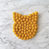 A handmade cat felt coaster rests on a marble surface, displaying its charming and whimsical design. This unique 4-inch wool felt cat coaster is crafted from tightly arranged eco-friendly felt balls, forming a distinct cat shape with subtle ears, adding a cozy touch to home decor.