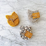 Three sets of handcrafted wool felt cat coasters are displayed on a marbled surface. Each coaster is approximately 4 inches in diameter and 0.5 inches thick, made from tightly arranged felt balls with subtle cat ear details. The sets come in mustard yellow, mixed gray, and solid light gray. Each set includes four coasters, bundled with jute twine and a branded tag that highlights their artisanal charm.